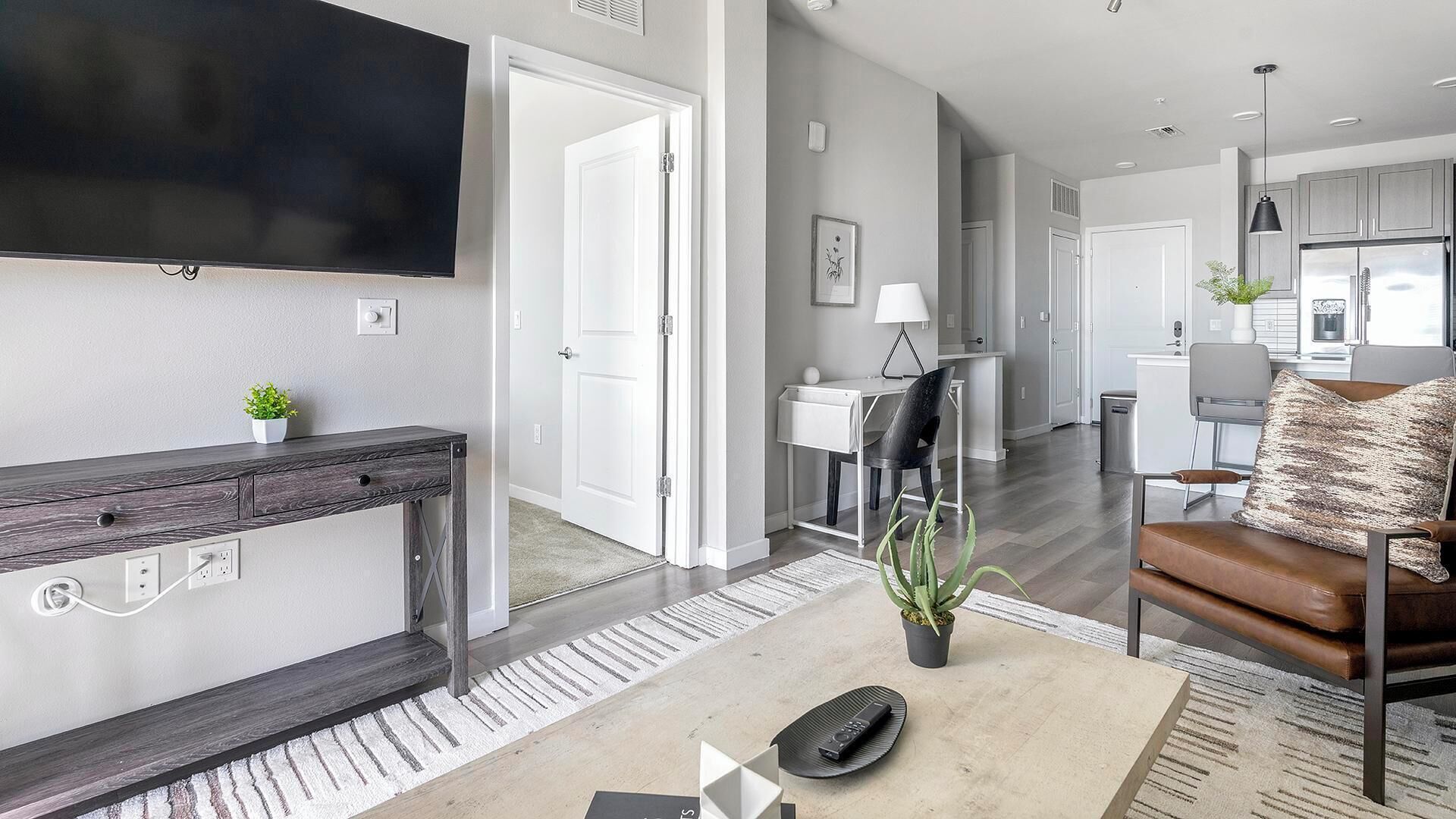 Een moderne woonkamer met een flatscreen-tv, een salontafel en een bank. Er is een deur die leidt naar een keuken en een eethoek.