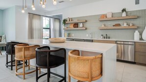 A modern kitchen with a large island, bar stools, pendant lighting, and built-in shelving. - Landing | Amazing 1BD, Gym, Game Room (Longboat Key)
