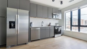 Uma cozinha moderna com armários cinza, uma geladeira de aço inoxidável, um micro-ondas e uma janela grande com vista para edifícios.