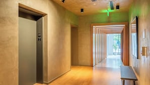 A modern hallway with a wooden floor, a wall-mounted desk, and a door marked with an elevator sign. - Landing | Modern 1BD, Rooftop Lounge, Pool (Durham)