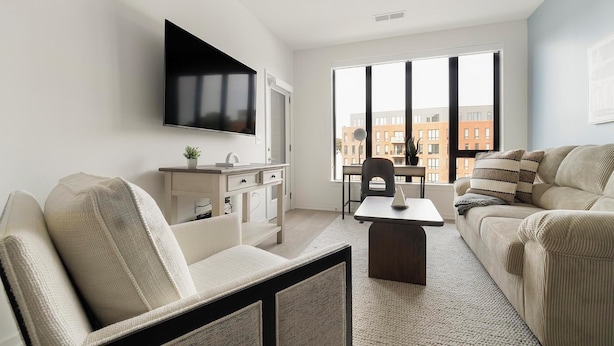 A modern living room with a flat-screen TV mounted on the wall, a sofa, a coffee table, and a desk with a chair. - Landing | Modern 1BD, Rooftop Lounge, Pool (Durham)