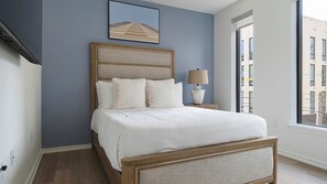 Une chambre à coucher avec un lit, une table de chevet, une lampe, un tableau encadré au mur et une fenêtre.