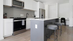 A modern kitchen with white cabinetry, a stainless steel oven, a microwave, and a refrigerator. There are two bar stools and a small island with a sink. - Landing | Spectacular 3BD, Pool, Gym (Durham)