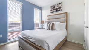A bedroom with a large bed, two windows with blinds, a bedside table with a lamp, and a wall-mounted artwork. - Landing | Spectacular 2BD, Gym, Rooftop Lounge (Durham)