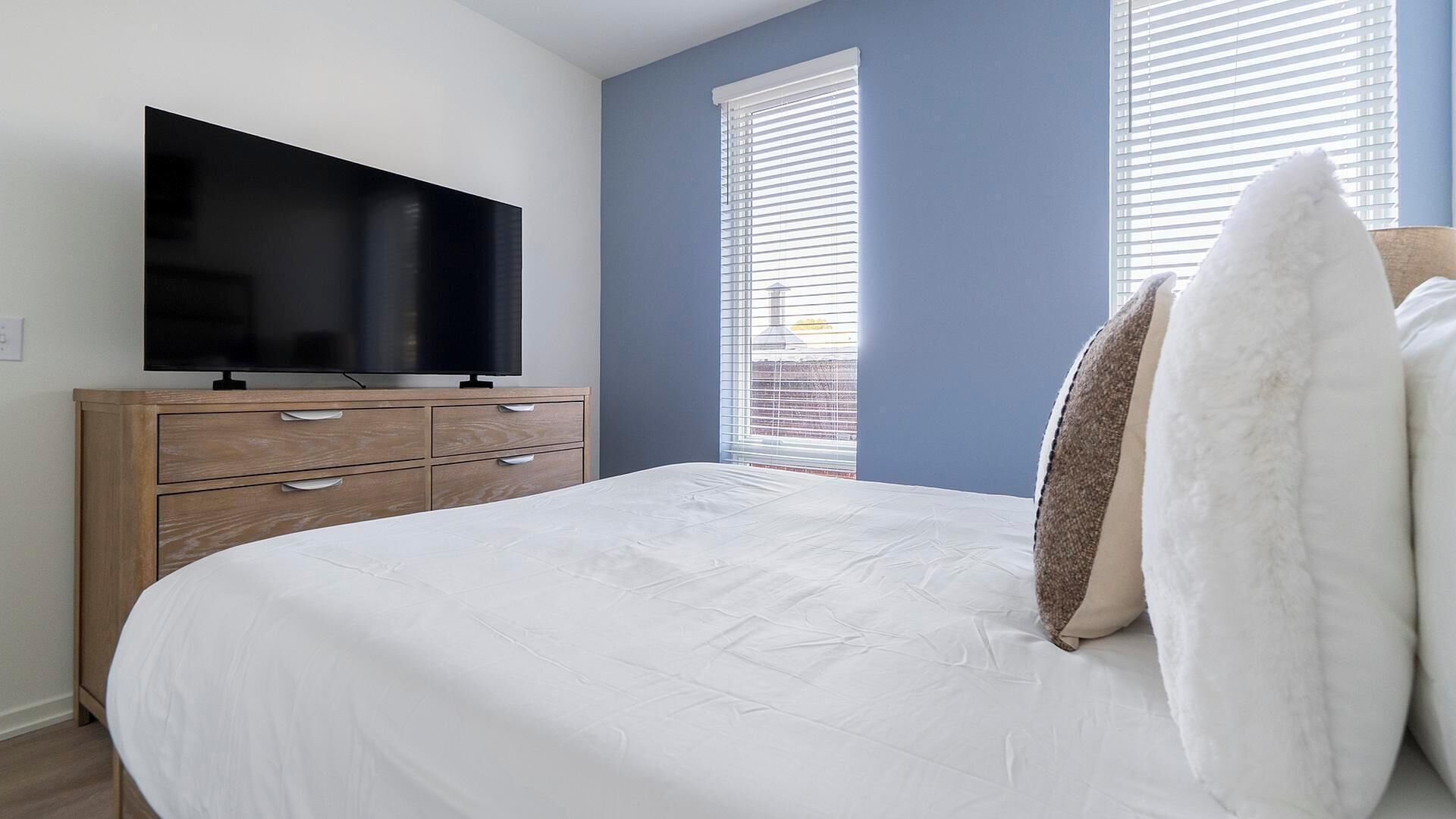 Un dormitorio con una cama, un televisor sobre una cómoda de madera y dos ventanas con persianas.