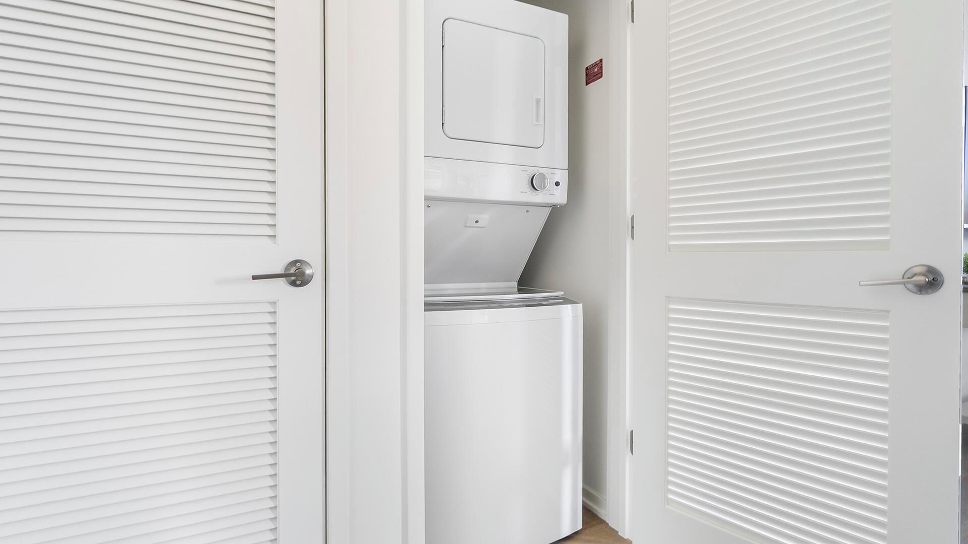 Une pièce blanche équipée d’un lave-linge et d’un sèche-linge intégrés, d’une porte avec une poignée et d’une fenêtre avec des stores horizontaux.