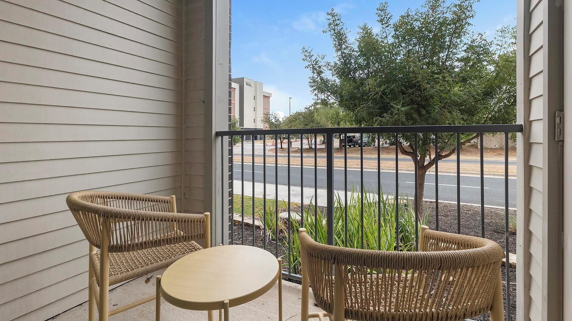 A balcony with two wicker chairs and a small table, overlooking a street with a tree and buildings in the background.