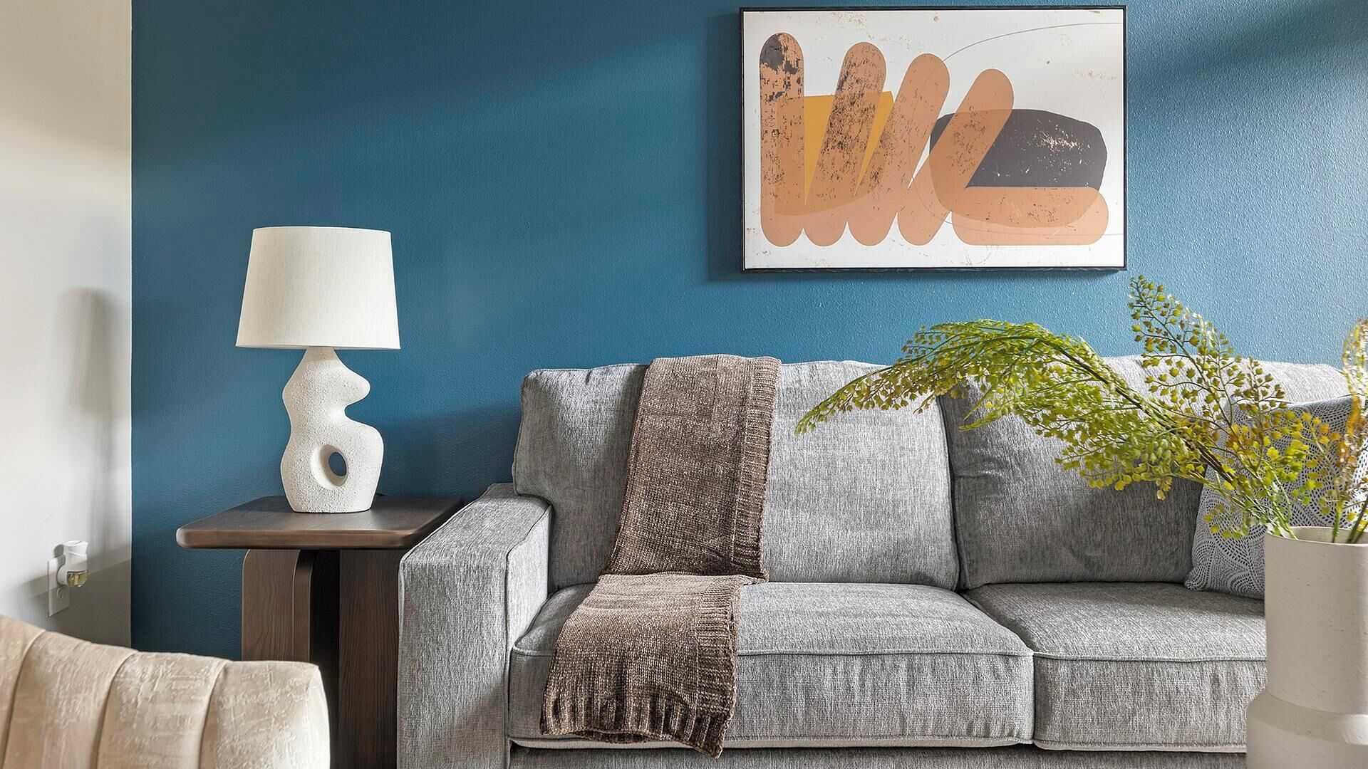 Sala de estar com parede azul, sofá cinza, luminária branca e uma obra de arte abstrata emoldurada.