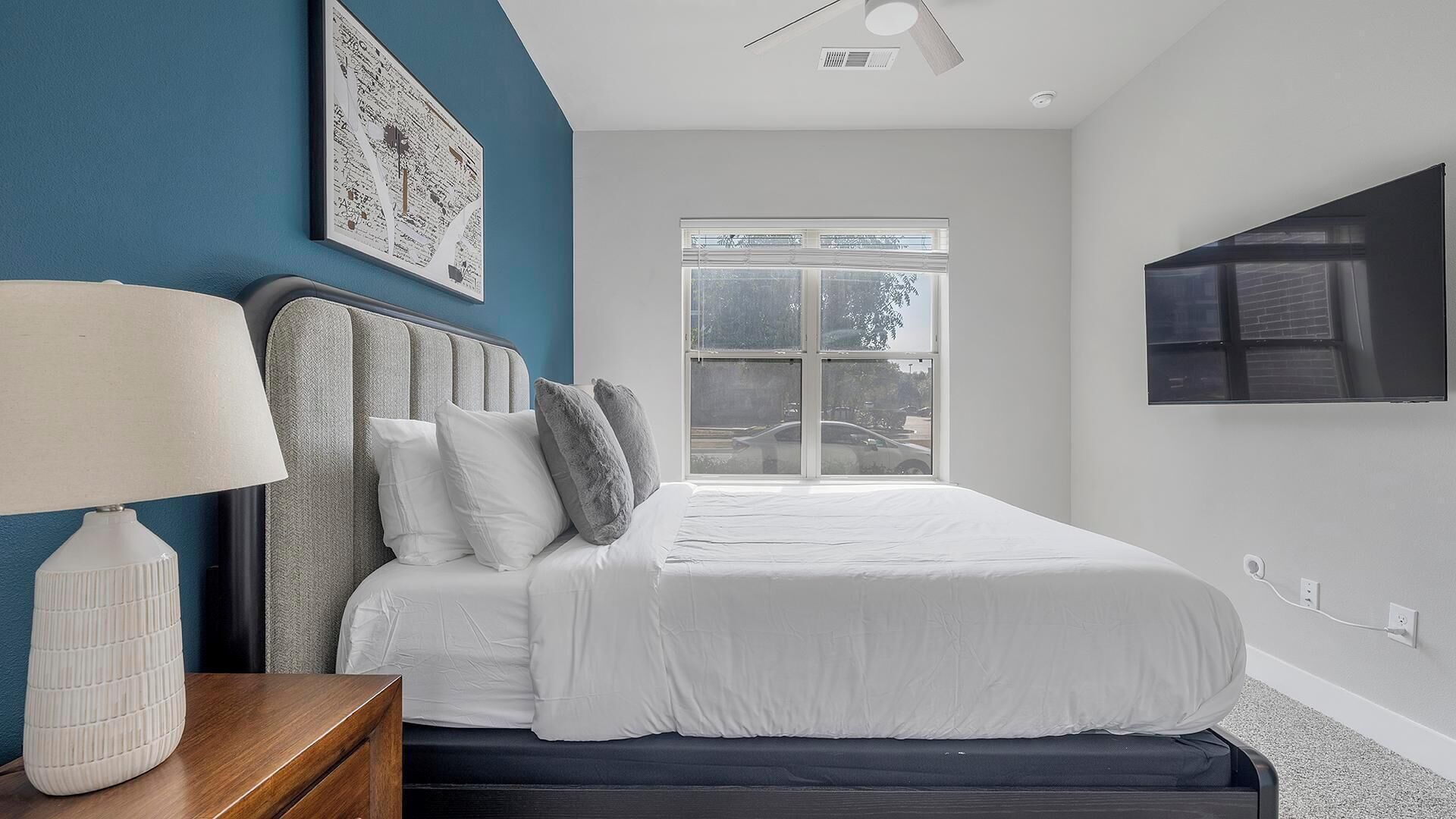A modern bedroom with a bed, a nightstand, a lamp, a large window, and a wall-mounted TV.