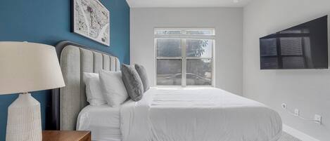 A modern bedroom with a bed, a nightstand, a lamp, a large window, and a wall-mounted TV.