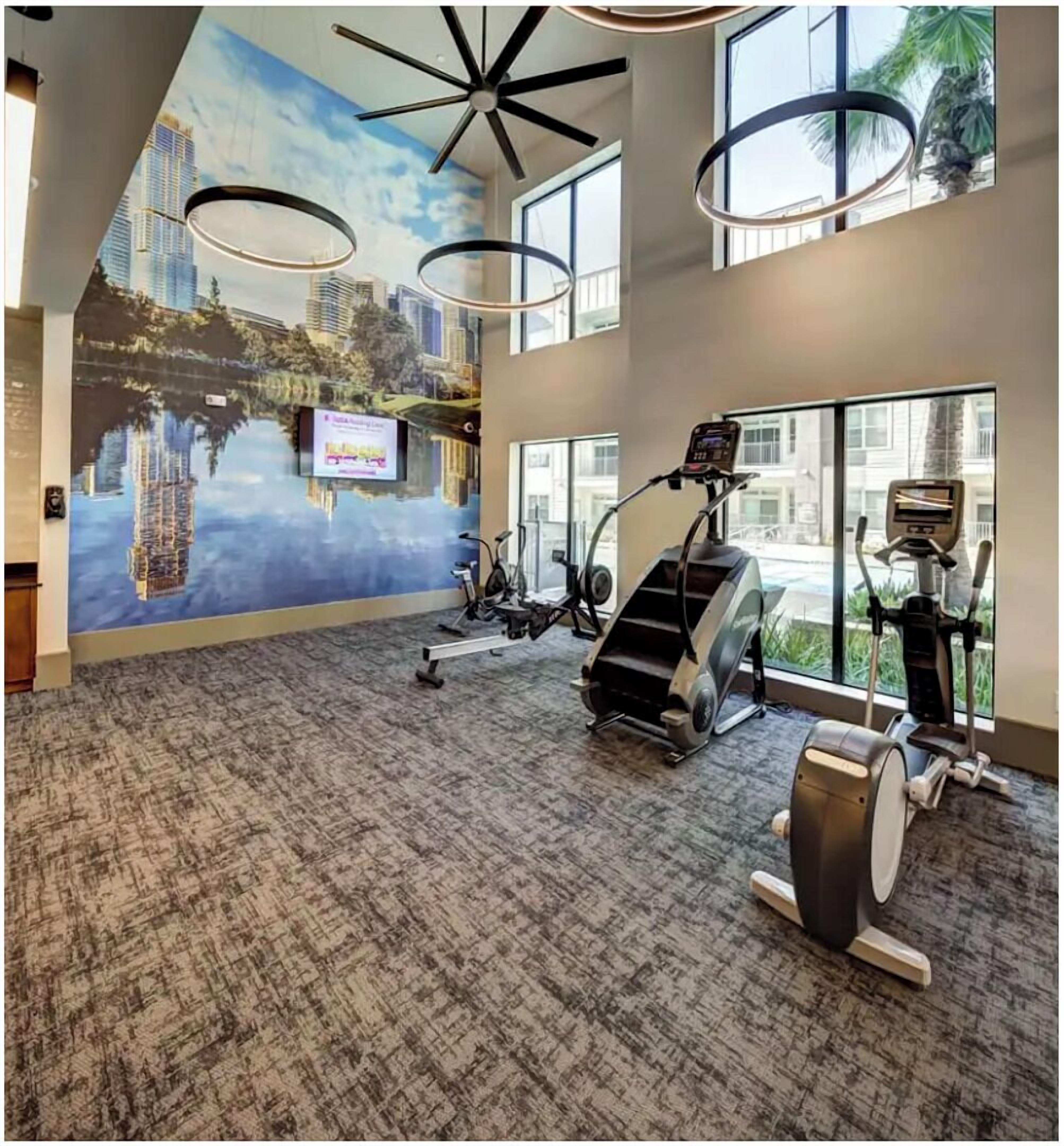 Uma academia ampla com equipamentos de cardio, um mural com vista para a cidade e grandes janelas com vista para o exterior.