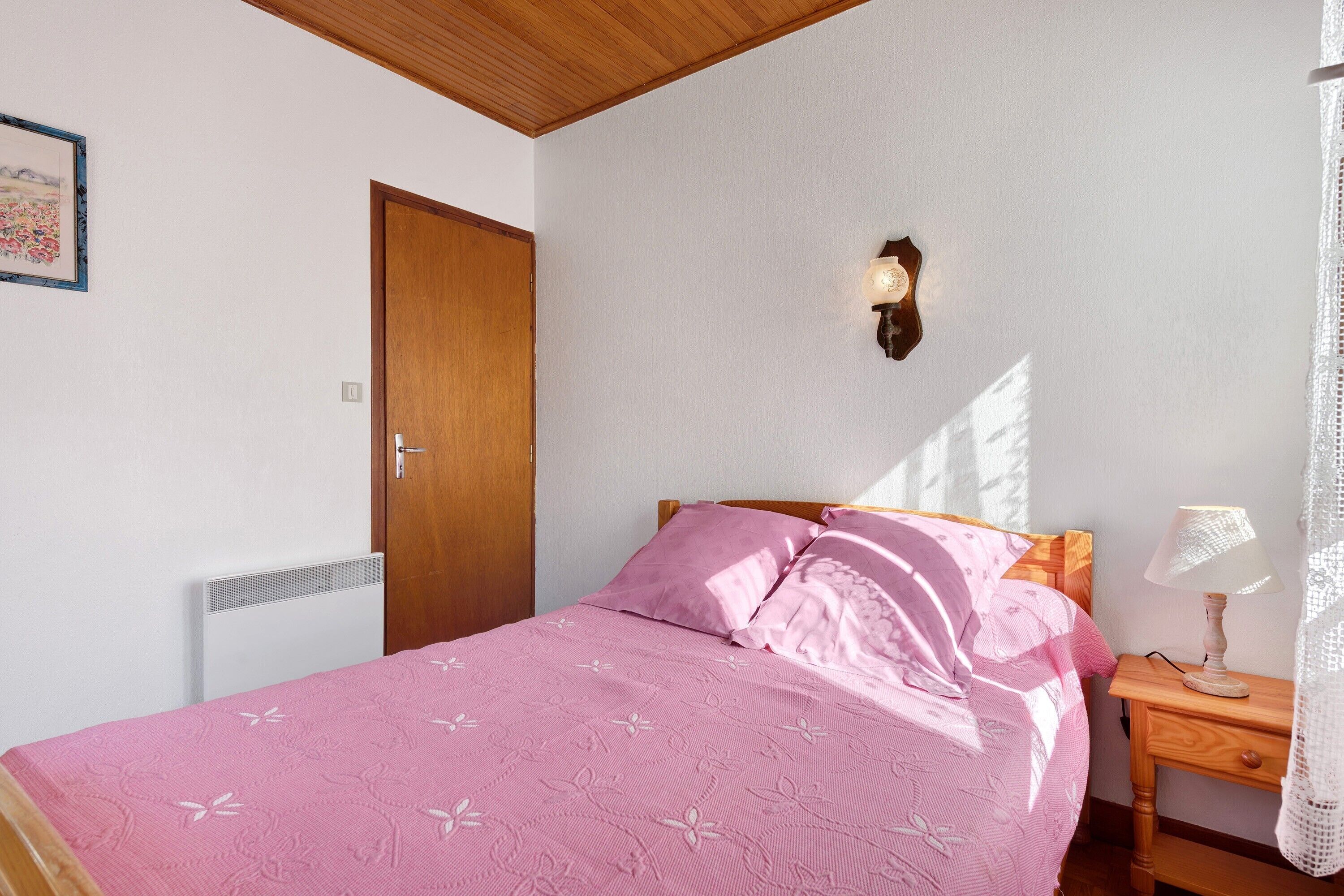 A bedroom with a bed, a nightstand, a wall-mounted lamp, a door, and a radiator.