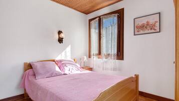 Een slaapkamer met een houten bed, een nachtkastje, een lamp, een raam met kantgordijnen en een ingelijst schilderij aan de muur.