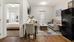 A compact living space with a bed, a dining area with a table and chairs, a sofa, a black refrigerator, and a TV mounted on the wall.