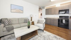 A modern living room with a sofa, a coffee table, and a potted plant. There is a kitchen area with cabinets and a microwave. - Landing | Incredible 2BD, Pool, Courtyard (Richmond)