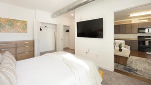 A bedroom with a bed, a wardrobe, a flat-screen TV, and a kitchen visible through an open door. - Landing | Incredible 2BD, Clubhouse, Courtyard (Richmond)