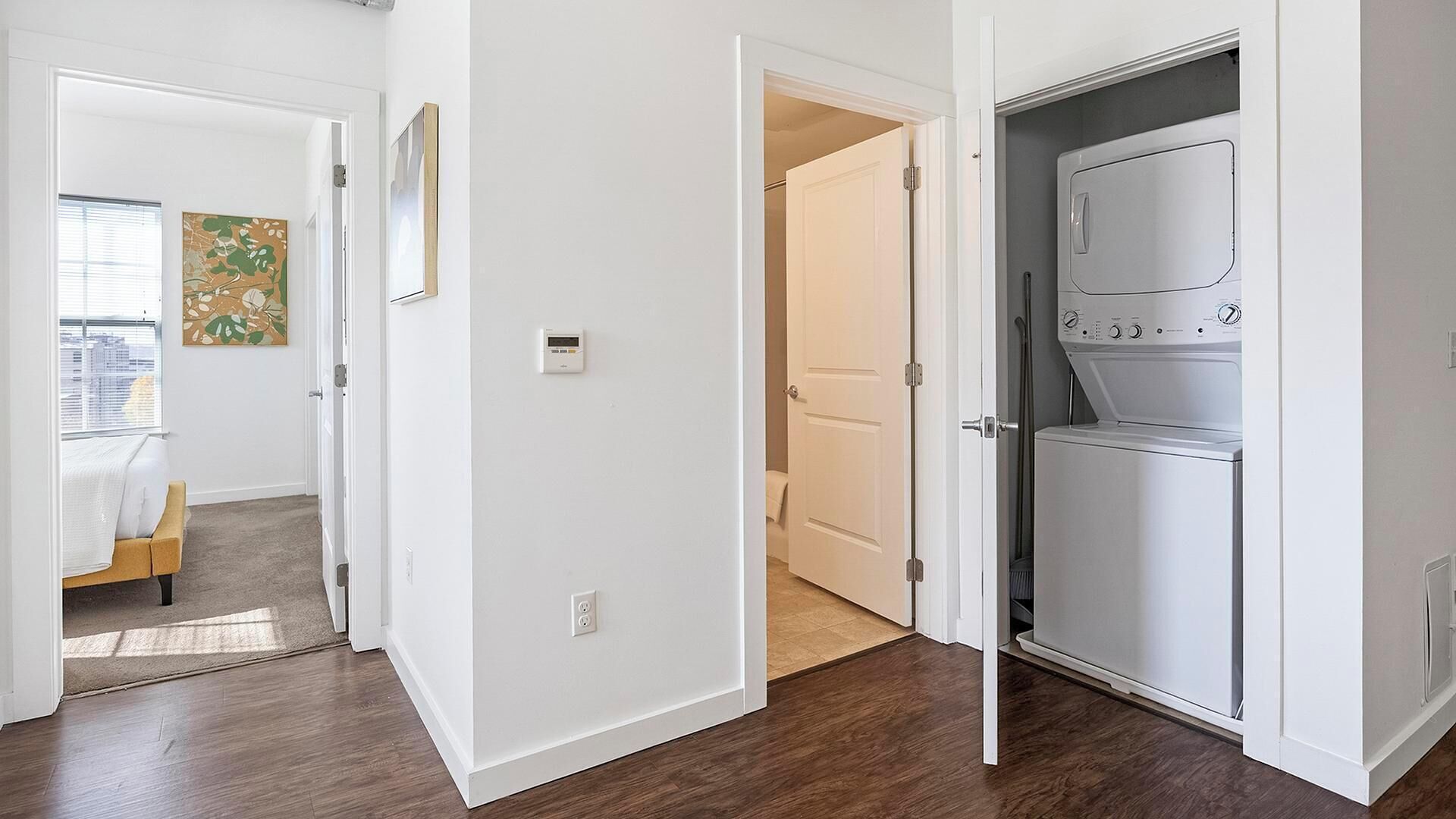 Eine moderne Wohnung mit Holzboden, einer weißen Tür zu einem Hauswirtschaftsraum mit Waschmaschine sowie einem Schlafzimmer mit Bett und Fenster.