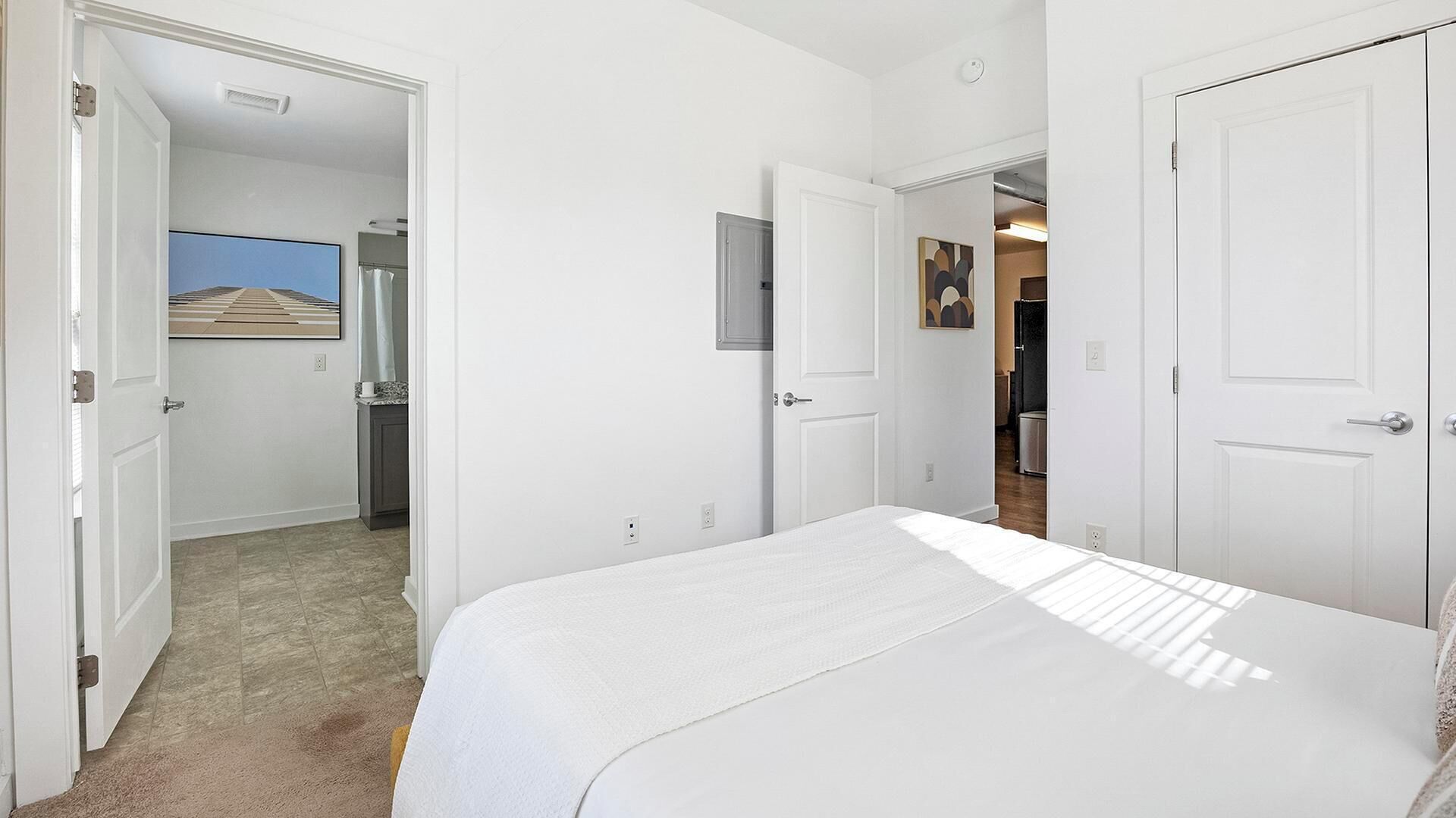 A hotel room with a bed, a door leading to a bathroom, and a hallway with a kitchen area visible through an open door.