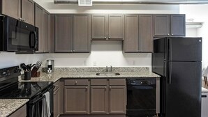 A modern kitchen with dark wood cabinets, a black refrigerator, a stainless steel microwave, and a granite countertop. - Landing | Chic 2BD, Rooftop Lounge, Courtyard (Richmond)