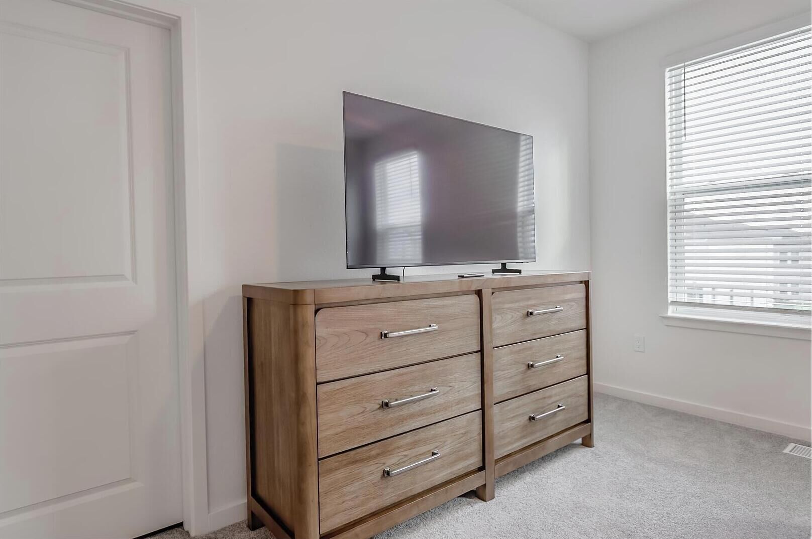 A room with a wooden dresser, a flat-screen TV on top, and a window with blinds.