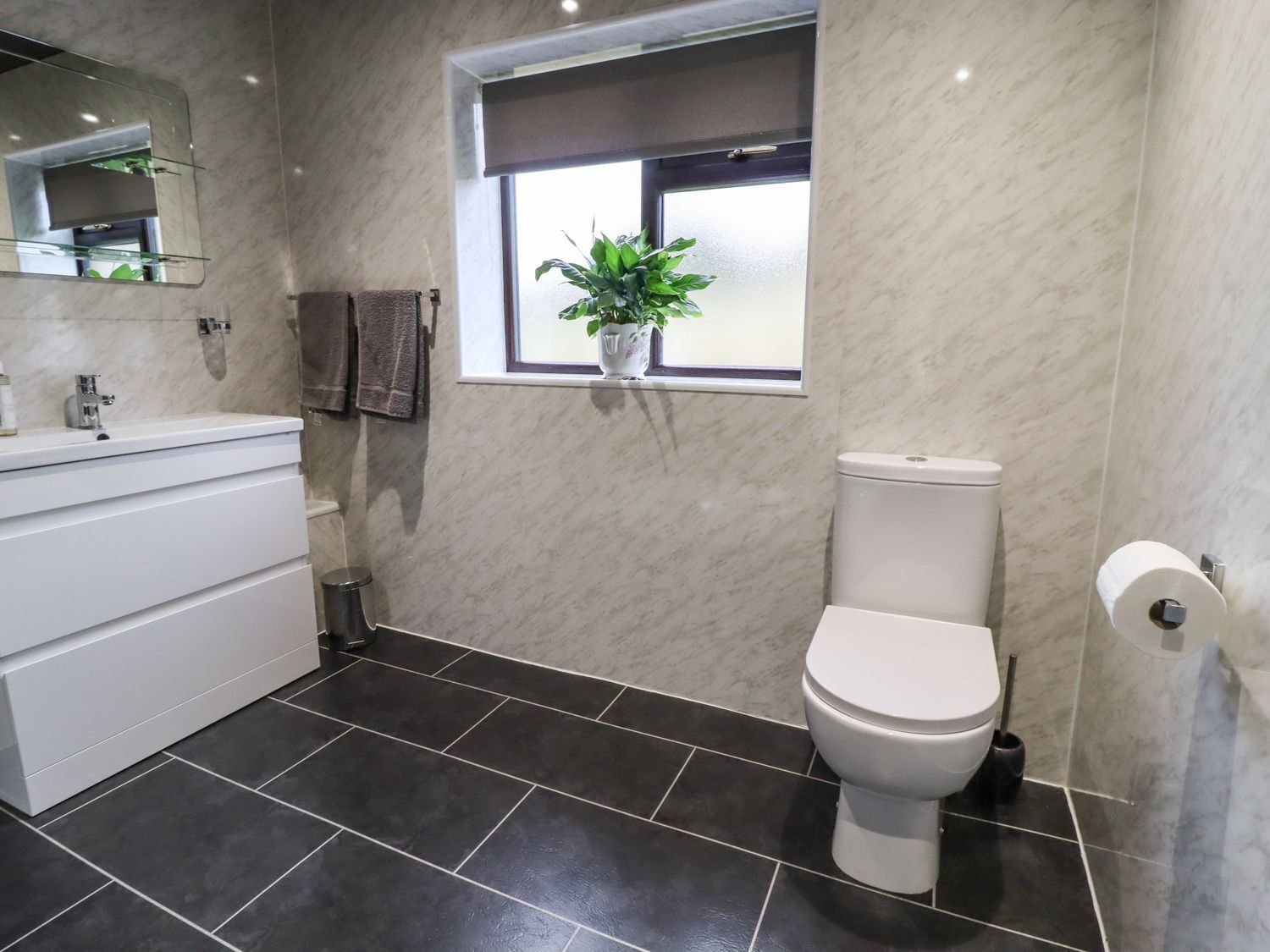 Une salle de bain moderne avec des toilettes blanches, un sol carrelé foncé, un meuble-lavabo blanc avec un miroir et une fenêtre agrémentée d’une plante.