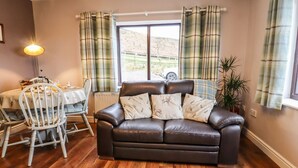 Ein Wohnzimmer mit einer Ledersofa, Karomustert-Vorhängen, einem Esstisch mit Stühlen und Blick auf eine ländliche Landschaft.