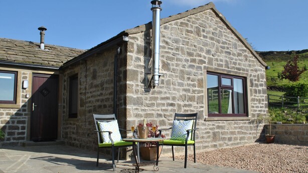 Ein Steinhaus mit Schornstein, zwei Fenstern und einer Terrasse mit zwei Stühlen und einem Tisch.