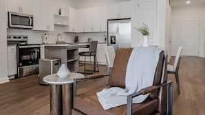 A modern kitchen with white cabinets, stainless steel appliances, and a dining area with a brown leather chair and a small table. - Landing | Stylish 3BD, Gym (Four Corners)