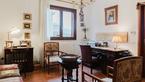 Ein Zimmer mit einem Bett, einem Schreibtisch, einem Sessel, einem Tisch, einer Chandelier und einem Fenster mit Vorhängen.
