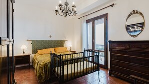 Ein Schlafzimmer mit Bett, Kommode, Spiegel, einer Chandelierleuchtre und einem Vorhangfenster.