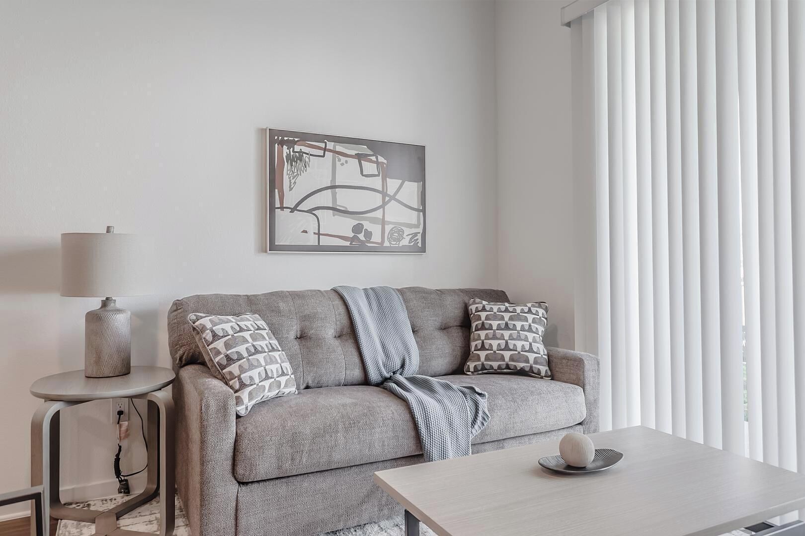 Una sala de estar moderna con un sofá gris, cojines estampados, una mesita de noche con una lámpara y una obra de arte abstracta enmarcada en la pared.