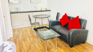 A modern living area with a glass coffee table, a black leather sofa with red cushions, a small round table, and a kitchen in the background. - City Sparrow Apartments (London)