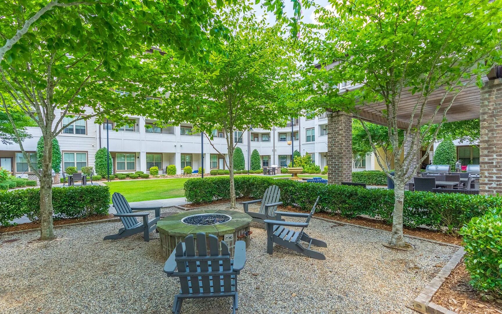 An outdoor seating area with a fire pit, surrounded by gravel and trees, with a building in the background.