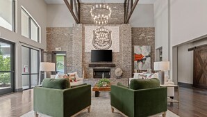 A spacious living room with a brick fireplace, two green armchairs, a coffee table, and a large TV mounted above the fireplace. - Landing | Spectacular 1BD, Gym, Courtyard (Little Rock)