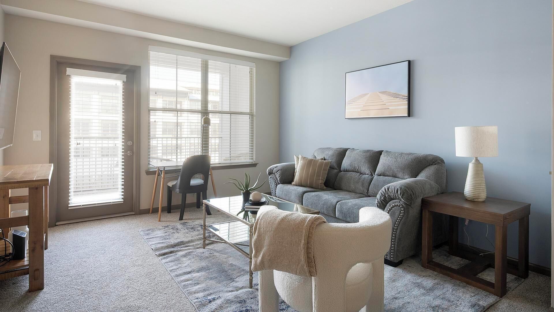 A living room with a grey sofa, a coffee table, a lamp, and a chair. There is a window with blinds, a picture on the wall, and a small table with a lamp.