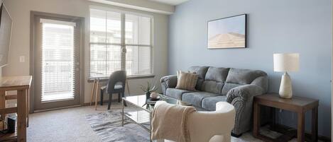 A living room with a grey sofa, a coffee table, a lamp, and a chair. There is a window with blinds, a picture on the wall, and a small table with a lamp.