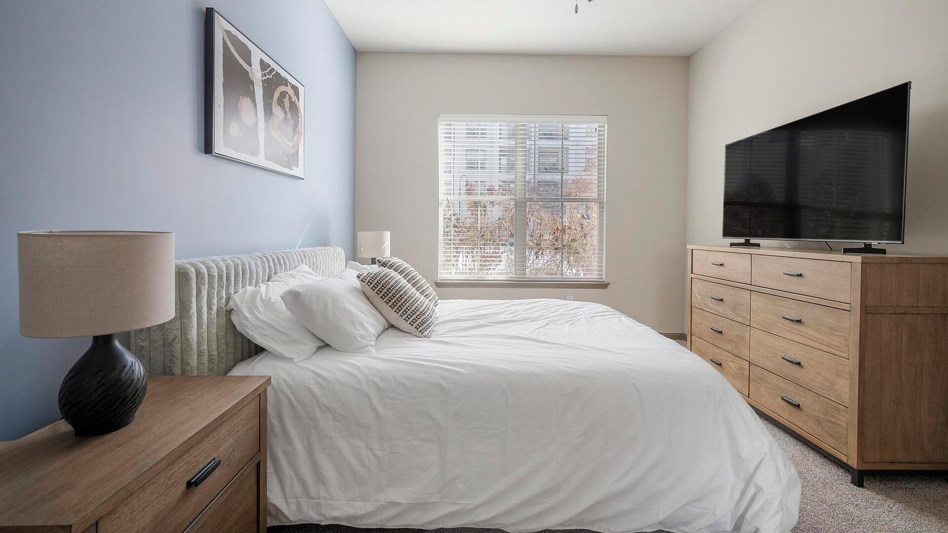 Quarto com cama, mesa de cabeceira, cômoda, televisão e janela com persianas.