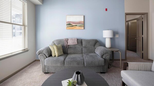 A living room with a grey sofa, a small table with a vase, a lamp, and a painting on the wall. - Landing | Amazing 2BD, Yoga Studio, Clubhouse (Little Rock)