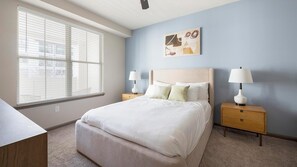 A bedroom with a bed, two bedside tables, a nightstand, a wall-mounted lamp, and a window with blinds. - Landing | Amazing 2BD, Yoga Studio, Clubhouse (Little Rock)