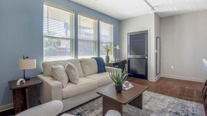 Un salon moderne avec un canapé beige, une table basse et une lampe. De grandes fenêtres sont équipées de stores, une porte dotée d’une poignée en métal et une plante ornent la table basse.