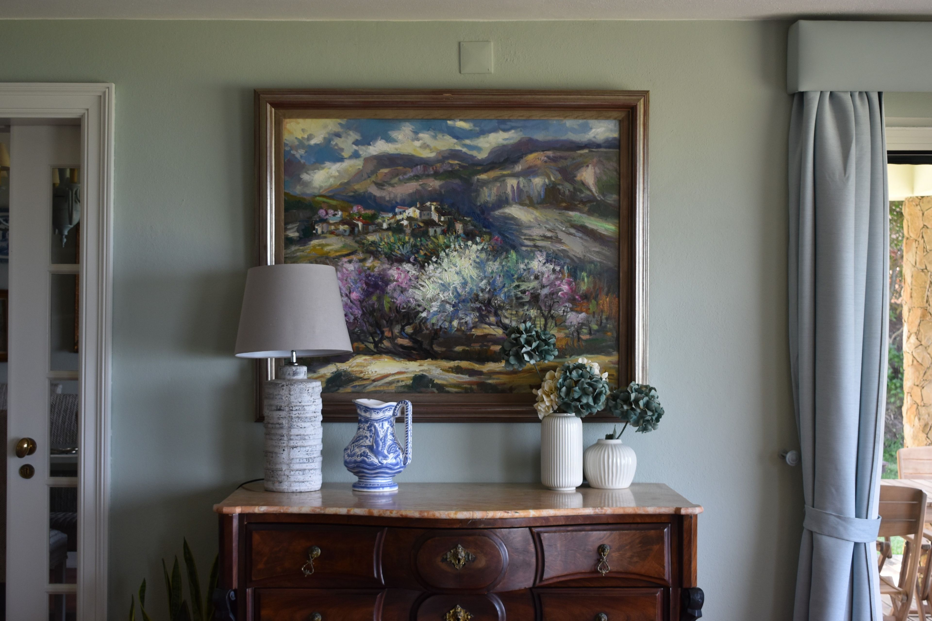 A room with a painting on the wall, a lamp, and a dresser with decorative items.