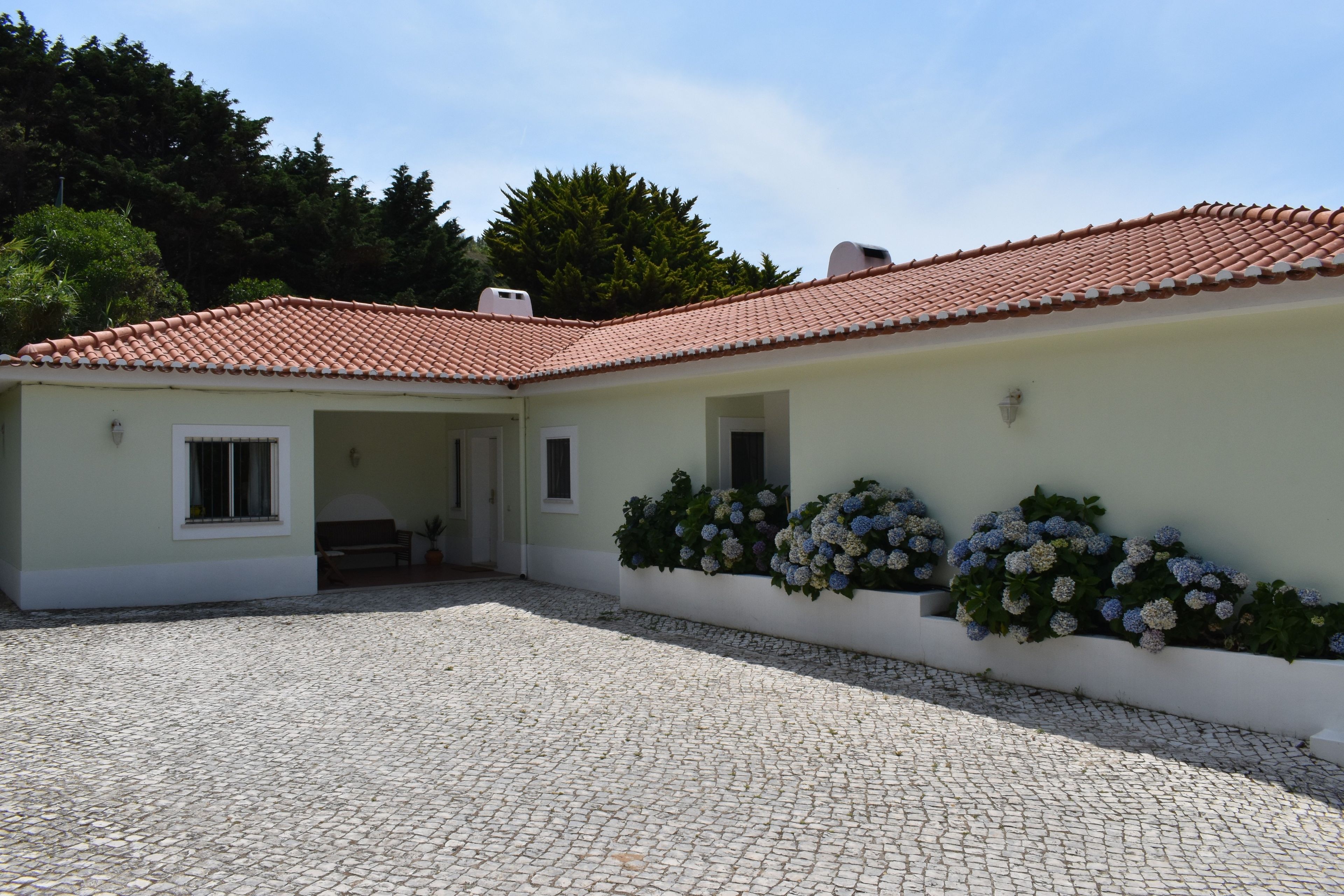 A building with a tiled roof, a paved courtyard, and a flower bed with hydrangeas.