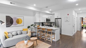 Una cocina moderna con armarios blancos, una isla central con taburetes de bar y un área de comedor con sofá gris y mesa de madera redonda.