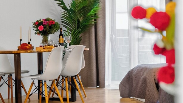 Ein modernes Wohnzimmer mit Essbereich, einem großen Fenster mit Vorhängen, einer Topfpflanze und einem Bett mit gesteppter Bettdecke.