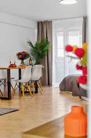 Een moderne woonkamer met een eethoek, een groot raam met gordijnen, een kamerplant en een bed met een gewatteerde spreideken.