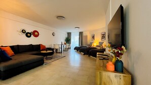 Ein modernes Wohnzimmer mit einer Couch, einem Couchtisch und einem Fernseher auf einem Holzboden. Im Hintergrund sind zwei Betten, eine Pflanze und ein Fenster mit Vorhängen zu sehen.