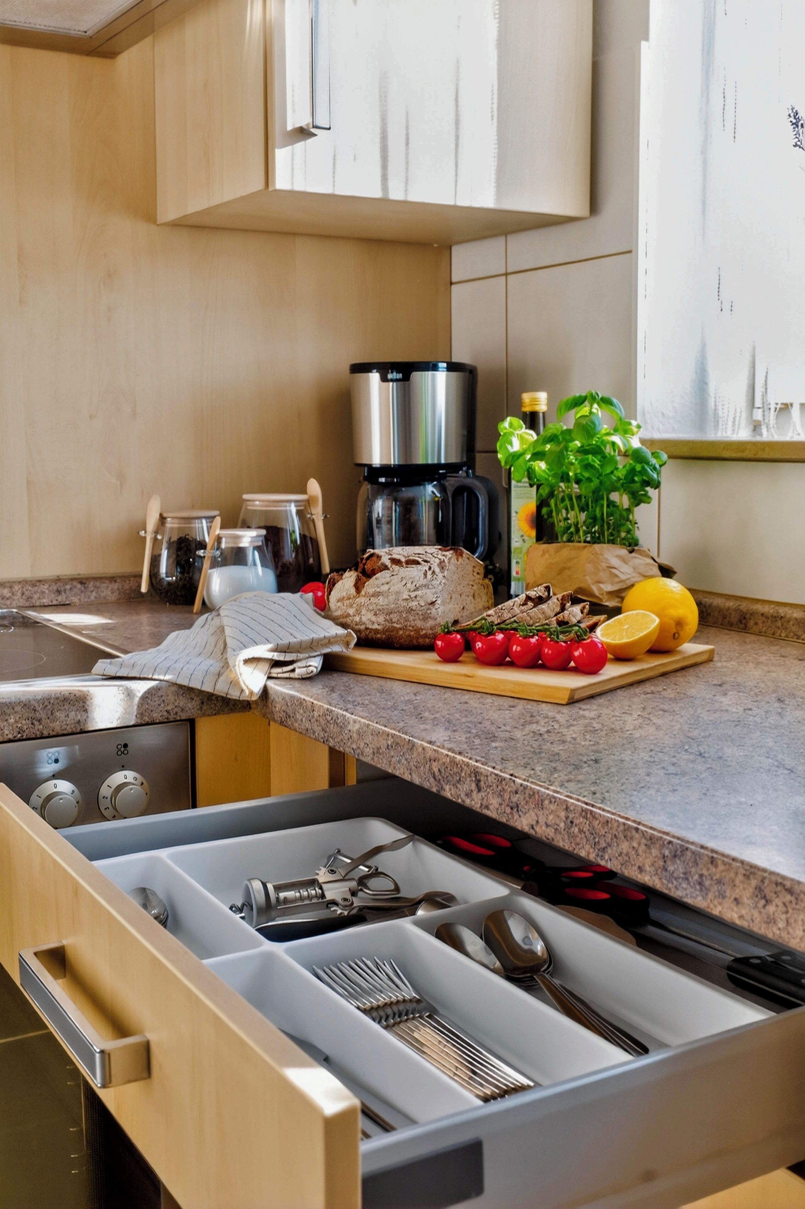 A kitchen with wooden cabinets, a stainless steel coffee maker, a cutting board with vegetables, and a drawer organizer.