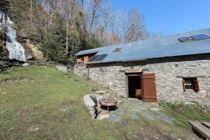 Un bâtiment rustique en pierre, doté d’un toit en métal, d’une porte en bois et d’un chemin en pierre menant à un espace barbecue.