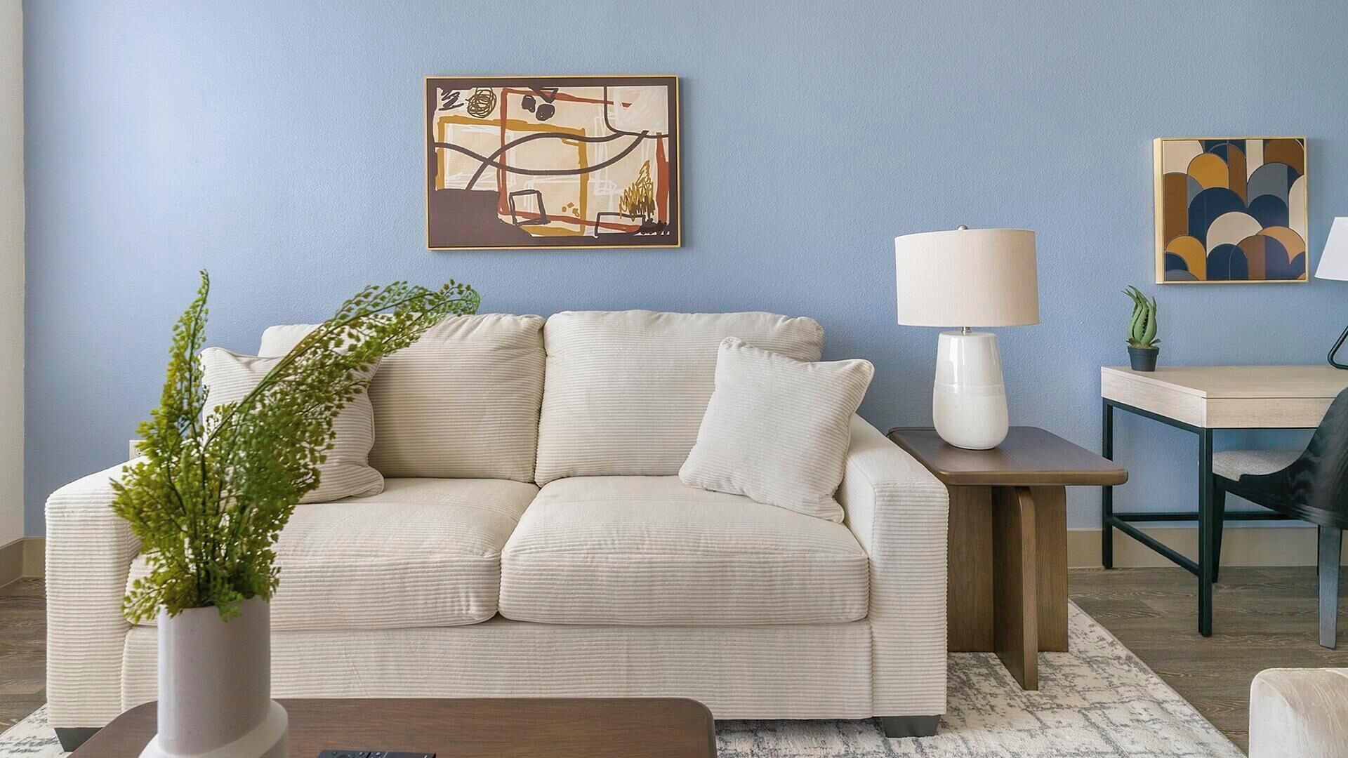 Una sala de estar con un sofá beige, una mesita de noche con lámpara, un escritorio pequeño con silla, y un cuadro abstracto enmarcado en la pared.