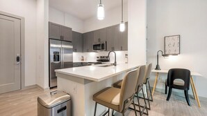 A modern kitchen with grey cabinets, stainless steel appliances, a white countertop, and bar stools. - Landing | Amazing 1BD, Gym, Game Room (Austin)